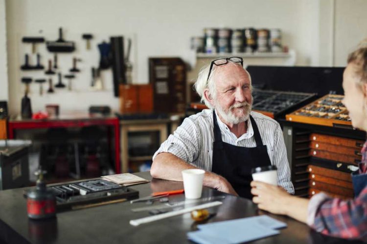 Senior craftsman sitting at table with young craftswoman and smiling in book arts workshop