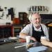 Senior craftsman sitting at table with young craftswoman and smiling in book arts workshop